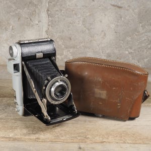 Peut inclure: Un appareil photo pliant Kodak vintage avec un corps gris et noir, un soufflet en accordéon noir et un objectif circulaire aux détails en métal argenté. À côté de l'appareil se trouve un étui de transport en cuir marron avec des coutures.