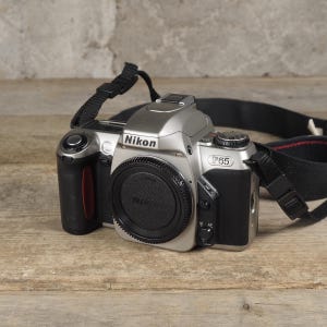 Puede incluir: Una cámara réflex de película de 35mm Nikon F65 en color plata y negro, con tapa de objetivo negra, empuñadura negra texturizada, dial de modo en la parte superior y una correa ajustable negra sujeta a los lados del cuerpo.