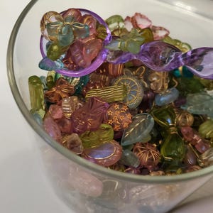 May include: A clear glass jar filled with an assortment of colorful beads. The beads are various shapes and sizes, including flowers, leaves, and geometric designs. The colors include pink, green, purple, and gold. A purple spoon rests on top of the beads.