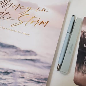 May include: A study book titled Mercy in the Storm: A Study on the Book of Jonah with a cover featuring a stormy ocean scene, a light blue pen, a light blue highlighter with the word Wisdom, and a bookmark with the text Be Still and Know That I Am God.