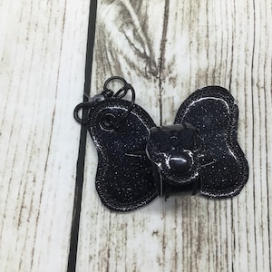 May include: Black glitter bow with a black metal ring and a black metal clasp. The bow is on a light wood background.