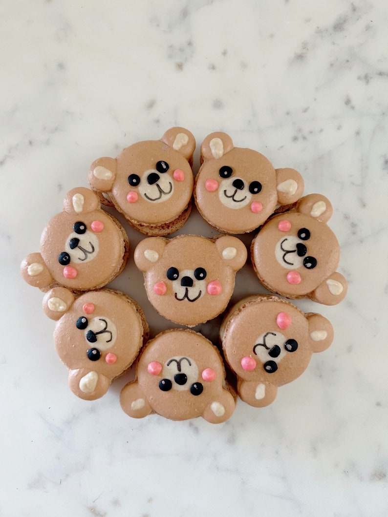 K&ouml;nnte beinhalten: Acht braune B&auml;ren-Macarons mit rosa Wangen und schwarzen Augen. Die Macarons sind in einem Kreis auf einer wei&szlig;en Marmorfl&auml;che angeordnet.