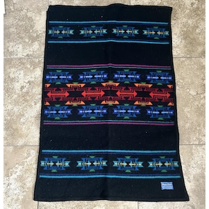 May include: A black rectangular blanket featuring horizontal bands of geometric patterns in shades of red, orange, blue, green, and purple. A small blue rectangular tag with white text is visible in the bottom right corner of the blanket.