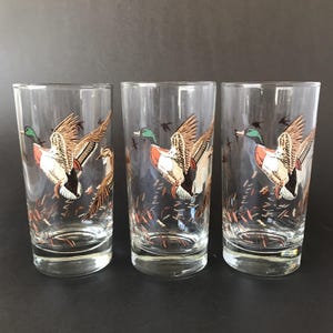 May include: Three clear drinking glasses with a detailed illustration of a mallard duck in flight. The ducks have brown, white, and orange feathers, with green heads. The glasses have a vintage aesthetic.