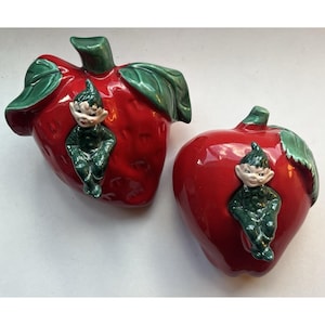 May include: Two glossy ceramic figurines shaped like bright red strawberries with green leaves. Each strawberry features a small, smiling elf figure with a green outfit and pointed hat attached to the front, creating a whimsical decorative set.