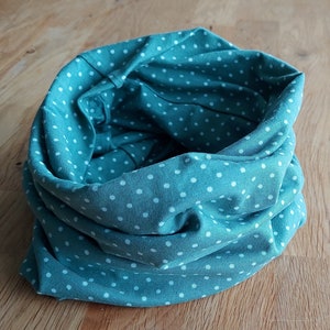 Könnte beinhalten: Ein türkisfarbener Stoff-Halswärmer mit weißen Punkten. Der Halswärmer ist aufgerollt und liegt auf einer Holzoberfläche.
