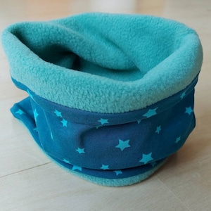 Könnte beinhalten: Ein türkisfarbener Fleece-Halswärmer mit einem türkisfarbenen und weißen Sternenmuster. Der Halswärmer ist in der Hälfte gefaltet und liegt auf einer hellbraunen Holzoberfläche.