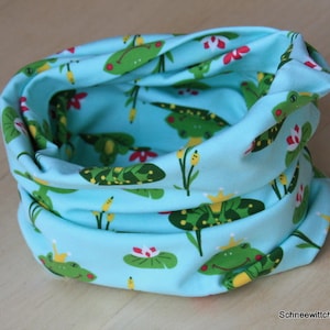 Könnte beinhalten: Ein hellblauer Stoff-Halswärmer mit einem sich wiederholenden Muster aus grünen Fröschen auf Seerosen. Die Frösche tragen Kronen.