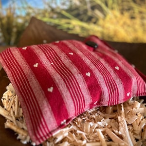 Könnte beinhalten: Ein rot-weiß gestreiftes Kissen mit kleinen weißen Herzen, das auf Holzspänen liegt. Das Kissen hat einen Knopfverschluss und ist wahrscheinlich ein dekorativer oder therapeutischer Artikel.