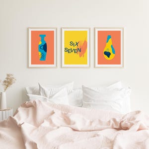 Puede incluir: Tres impresiones artísticas enmarcadas con diseños abstractos. Las impresiones presentan un jarrón, las palabras "Six Seven" y una forma de lágrima. Los fondos son naranja y amarillo, con acentos azules y amarillos. Las impresiones se muestran sobre una cama.