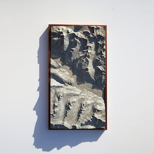 May include: A 3D relief map of a mountainous region, framed in a dark wood frame. The map is made of a light gray material and shows the contours of the mountains in detail.