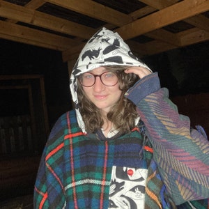 Könnte beinhalten: Eine Person trägt einen Kapuzenpullover mit schwarz-weißem Muster und darüber einen karierten Poncho. Der Poncho hat eine große Tasche mit einem schwarz-weißen Grafikdesign.