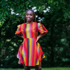 May include: A woman wearing a colorful, patterned dress with a short, flared skirt. The dress has a geometric design in shades of red, yellow, blue, and black. She is standing on a paved surface with green trees in the background.