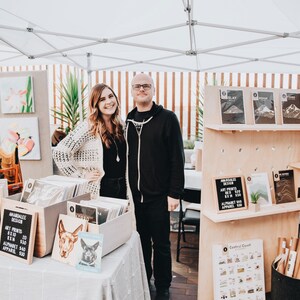 May include: A couple stands in front of their art and apparel booth at a market. The booth features a variety of items, including art prints, apparel, and a sign that reads "Remarkable Design". Art Prints: $10-$15, Apparel: $20-$40.
