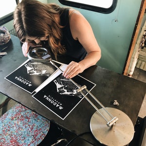 May include: Two black and white prints of a mountain range with the text "MADONNA MOUNTAIN" printed vertically on each print. A person is using a magnifying glass to examine the prints.