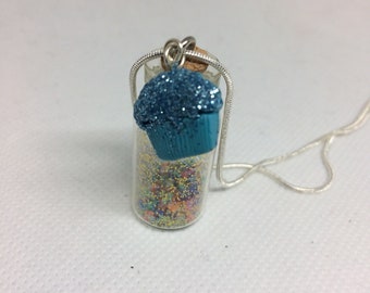 Könnte beinhalten: Eine Miniatur-Cupcake-Anhänger-Halskette. Der Anhänger zeigt eine kleine Glasflasche gefüllt mit bunten Streuseln, gekrönt von einem blauen Cupcake-Charm. Die Halskette hat eine silberne Kette und eine silberne Drahtöse.