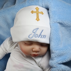 May include: A white baby hat with a gold cross and the name "John" embroidered in blue.