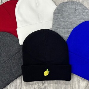 May include: Six knit beanies in various colors, including red, white, gray, black, and blue. One black beanie has a yellow lemon embroidered on the front.