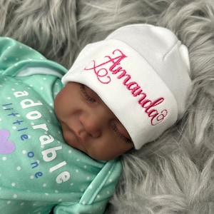 May include: White baby hat with the name "Amanda" embroidered in pink cursive lettering.
