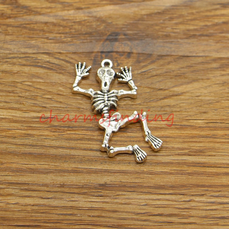 May include: Silver skeleton charm with a distressed finish. The skeleton is in a screaming pose with its arms raised and legs bent. The charm has a loop at the top for attaching to a necklace or bracelet.
