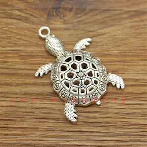 Puede incluir: Encanto de tortuga plateado con un diseño de caparazón calado detallado. El encanto tiene un bucle en la parte superior para sujetarlo a joyas u otras manualidades.
