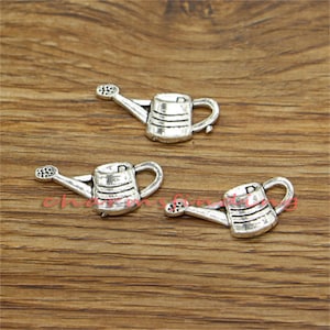 May include: Three silver-colored watering can charms. Each charm has a handle, a spout, and a detailed body with horizontal lines. The charms are arranged on a wooden surface.