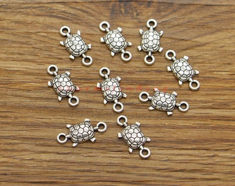 May include: Silver-toned turtle charms with two jump rings on each charm. The charms are scattered on a wooden surface.