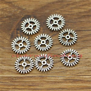 May include: Eight silver metal gear charms with a detailed design. The gears are all different sizes and have a slightly aged look.