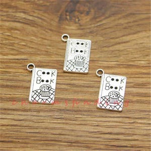 May include: Three silver charm pendants featuring the words "Cook Book" and a cupcake design. The charms are ideal for adding a touch of whimsy to jewellery or crafts.