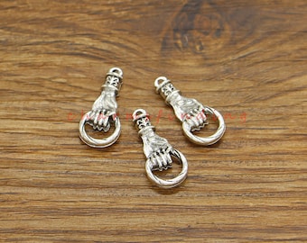 May include: Three silver-colored charms shaped like hands holding a ring. The charms have a detailed design, with the hand's fingers and the ring clearly visible. The background is a wooden surface.