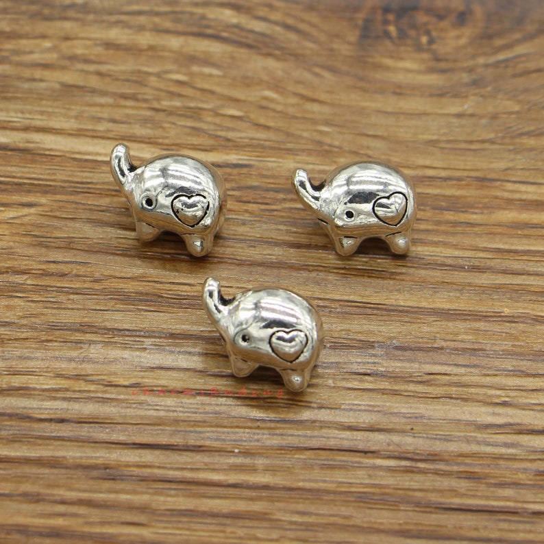 May include: Three silver elephant charms with a heart on each elephant's trunk. The charms are on a wooden surface.