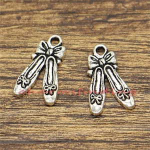 May include: Two silver-toned ballet slipper charms with bows. Each charm features a pair of ballet slippers with delicate bow details at the toe and top. The charms have a loop for attaching to jewelry or other crafts.