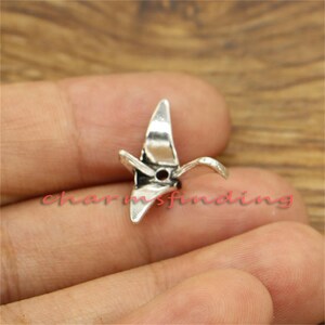 May include: Silver charm in the shape of a paper crane. The charm has a hole in the center for attaching to a bracelet or necklace.
