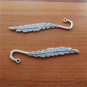 May include: Two silver feather-shaped bookmarks with a hook at the end. The bookmarks are on a wooden surface.