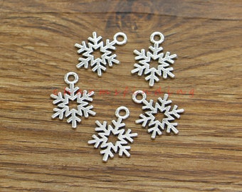 May include: Five silver-colored snowflake charms with a small loop at the top for stringing. Each charm has six points and a detailed, open center. The charms are displayed on a wooden surface.