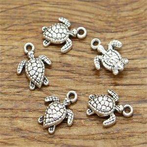 May include: Five silver turtle charms with a textured shell design and a loop for attaching to jewellery or other crafts. The charms are arranged on a wooden surface.