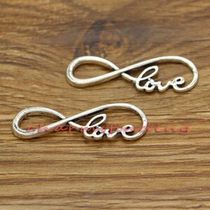 Könnte beinhalten: Zwei silberne Unendlichkeits-Anhänger mit dem Wort "love" in jedem Symbol. Die Anhänger liegen auf einer Holzoberfläche.