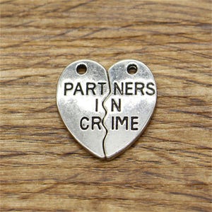 Könnte beinhalten: Silberfarbener herzförmiger Anhänger mit den Worten "Partners in Crime", die zwischen die beiden Hälften des Herzens aufgeteilt sind.