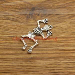 May include: A silver-toned skeleton charm featuring a detailed design. The skeleton is in a pose of fear, with its arms raised and its mouth open. The charm is ideal for adding a spooky touch to jewellery or crafts.