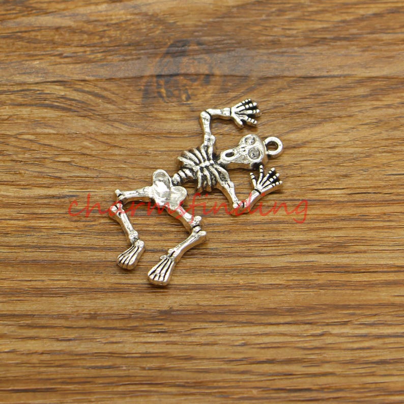 May include: A silver-toned skeleton charm featuring a detailed design. The skeleton is in a pose of fear, with its arms raised and its mouth open. The charm is ideal for adding a spooky touch to jewellery or crafts.