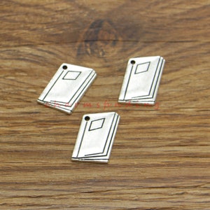 May include: Three silver-toned book charms with a hole at the top for attaching to a necklace or bracelet. The charms are detailed with a book spine and a closed book cover.