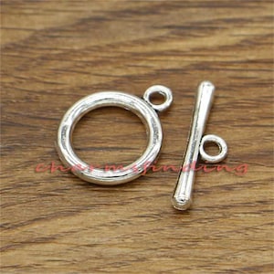 Puede incluir: Cierre de palanca de metal plateado con un anillo redondo y una barra recta. El cierre es un hallazgo común de joyería utilizado para collares, pulseras y otros proyectos de joyería.