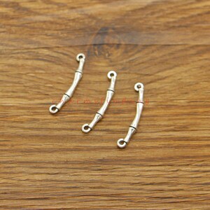 Op de afbeelding: Drie zilverkleurige metalen bedels in de vorm van bamboestokken met een lus aan elk uiteinde. De bedels zijn in een rij op een houten oppervlak gerangschikt.