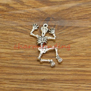 May include: Silver-toned skeleton charm featuring a detailed bone structure and a skull with a wide-open mouth. The skeleton is in a crouching position with its arms raised.