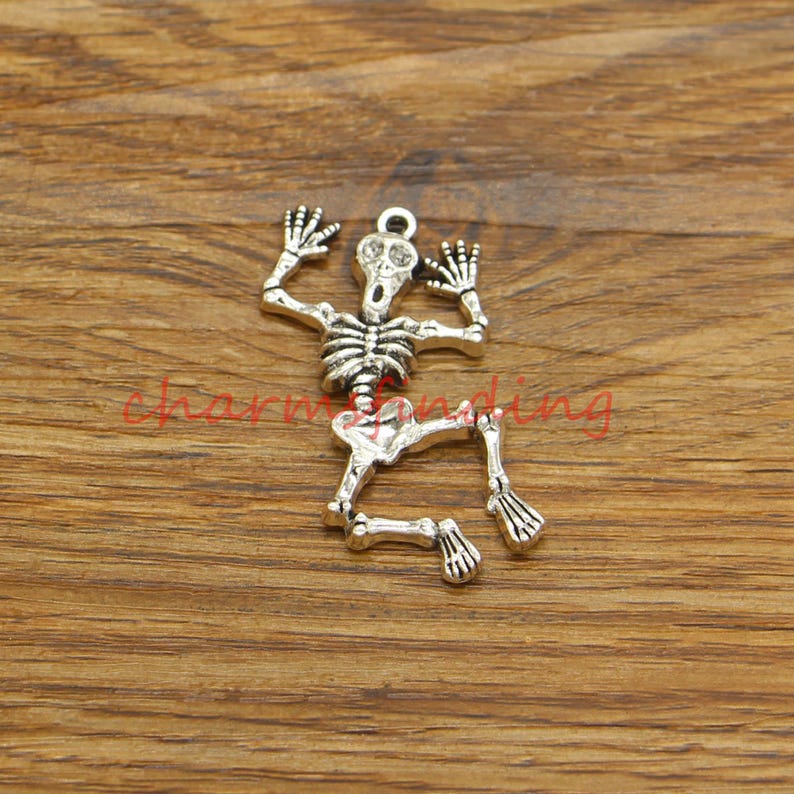May include: Silver-toned skeleton charm featuring a detailed bone structure and a skull with a wide-open mouth. The skeleton is in a crouching position with its arms raised.