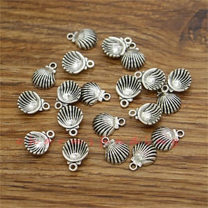 Puede incluir: Una colección de abalorios de metal plateados con forma de conchas marinas. Cada abalorio tiene un bucle en la parte superior para sujetarlo a joyas u otras manualidades.