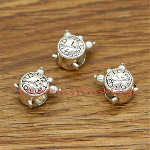 May include: Three silver charm beads in the shape of an alarm clock. Each bead has a raised clock face with black numbers and hands.