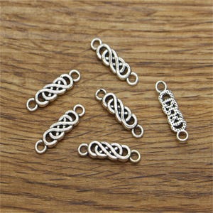 Pode incluir: Seis pingentes conectores de metal prateado com um design de nó intrincado. Cada pingente tem dois laços para prender a joias ou outros artesanatos.