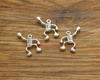 May include: Three silver-colored skeleton charms with outstretched arms and legs are displayed on a wooden surface. Each charm has a loop at the top for attaching to jewelry or other crafts. The charms are detailed with rib cages and rounded feet.