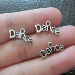 May include: Three silver charm pendants with the word "Dance" spelled out on each. The charms are on a hand, ready to be added to a jewellery project.